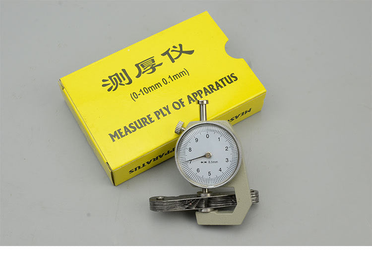 A dial thickness gauge with a silver body and dial, placed on a grey surface, with a yellow box in the background having Chinese characters printed on it.