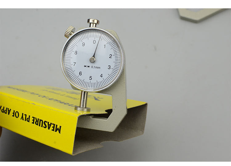 A dial thickness gauge with a silver body and dial, placed on a grey surface, with a yellow box in the background having Chinese characters printed on it.