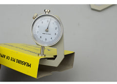 A dial thickness gauge with a silver body and dial, placed on a grey surface, with a yellow box in the background having Chinese characters printed on it.