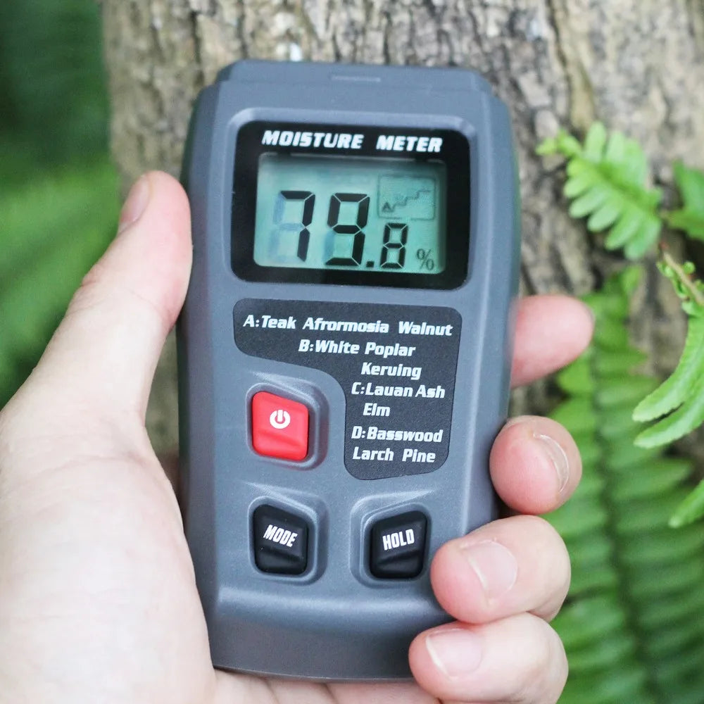 Hand holding a moisture meter against a tree with greenery in the background