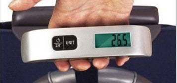 A hand holding a black luggage scale with a green LCD screen displaying weight, featuring a hook and button for operation.